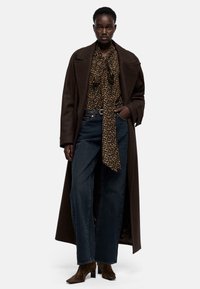 Lange bruine jas over een blouse met luipaardprint en een gestrikte halslijn, gecombineerd met donkerblauwe jeans met wijde pijpen en bruine enkellaarsjes.