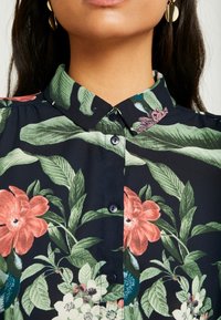 Blusa nera a fiori con vivaci fiori rosa e verdi, motivi a foglia, chiusura con bottone e design del colletto classico. Tessuto morbido.