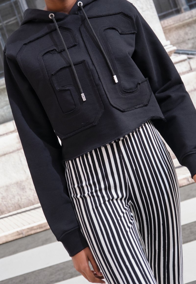 Zwarte cropped hoodie met grote, gestructureerde "GC" logo, met zilverkleurige hardware. Gepaard met zwart-wit verticaal gestreepte broeken.