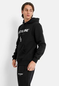 Felpa nera con cappuccio, stampa del logo, cappuccio con coulisse, tasca a marsupio, polsini e orlo a costine, indossata con pantaloni neri coordinati.
