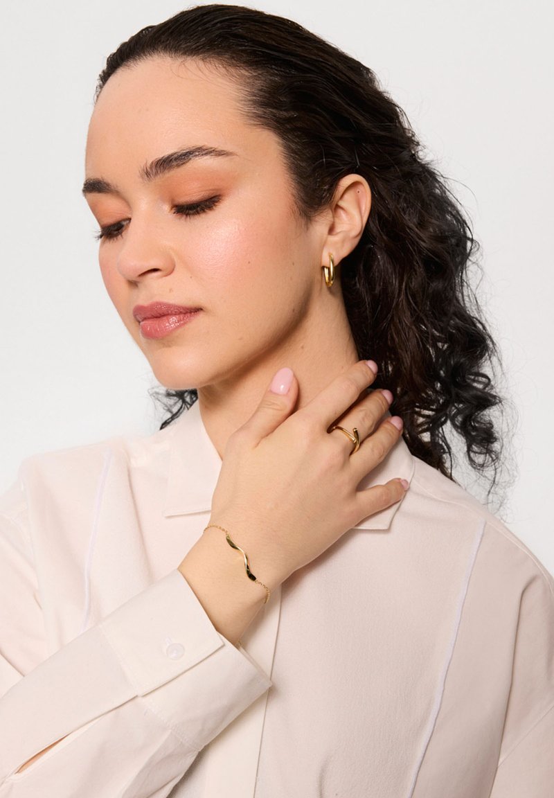 Biżuteria w złotym kolorze, w tym falowana bransoletka, pierścionek i kolczyki w kształcie kół, w połączeniu z jasnym bluzką z zapięciem na guziki przy mankietach.