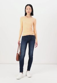Light orange ribbed long-sleeve top with scalloped neckline, paired with dark skinny jeans and white sneakers, holding a brown handbag.