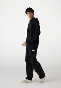 Sort hætte sweatshirt med kengurulomme og logo, parret med løse sorte bukser og hvide sneakers med beige detaljer.