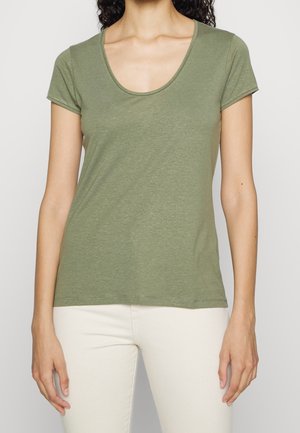 Kobieta w oliwkowozielonym t-shircie z krótkim rękawem i okrągłym dekoltem oraz kremowych spodniach, pokazana od szyi do bioder na jednolitym tle.
