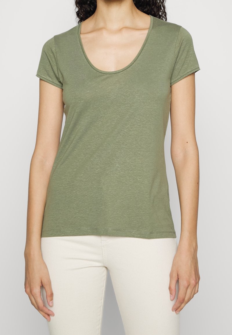 Femme portant un t-shirt vert olive à manches courtes avec encolure ronde et un pantalon blanc cassé, montrée du cou aux hanches sur un fond uni.