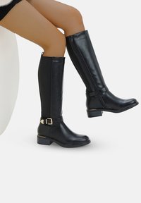 Botas negras hasta la rodilla, hechas de cuero suave, con cremallera lateral, detalle de hebilla y tacón bajo.