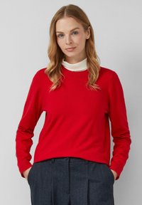 Czerwona, długodziełowa sweter z okrągłym dekoltem, noszony na białym golfie. Sweter ma ribbowe mankiety i gładką fakturę.