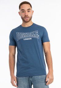 Mėlynas medvilninis marškinėliai su trumpomis rankovėmis, su baltu spauduotu logotipo "LONSDALE LONDON" per krūtinę. Lygi tekstūra, klasikinis kirpimas.