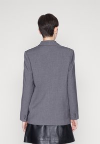 Blazer gris hecho de tela texturizada, con cuello con muescas, mangas largas con puños de botones y un corte ajustado, llevado sobre una falda negra.