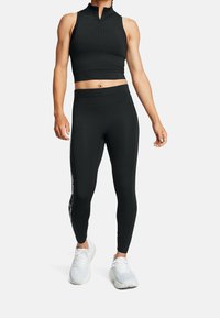 Czarny sportowy crop top z wysokim kołnierzem i zapięciem na zamek, w połączeniu z czarnymi legginsami z bocznym wzorem oraz białymi trampkami.