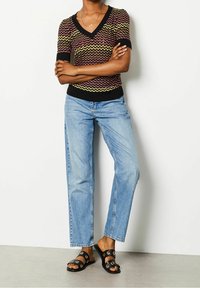 Pletený top s cik-cak vzorem v barvách tmavě červené, žluté a černé; spárován se světle modrými džíny s rovnými nohavicemi a černými skluznými sandály s kovovými akcenty.