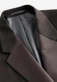 Blazer marrone scuro con un tessuto esterno texturizzato e interni grigi lisci. Tra le caratteristiche spiccano un colletto classico e sezioni di tessuto a contrasto.