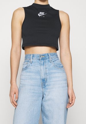 Femme portant un haut court sans manches noir Nike Air et un jean taille haute bleu clair, debout devant un fond uni.