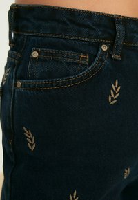 Jeans de un azul oscuro con un corte ajustado, con costuras doradas y patrones de hojas bordados en los bolsillos delanteros y laterales.
