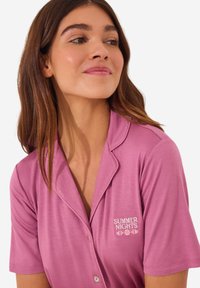 Roze kortemouwen pyjama top met een kraag, met "ZOMERNACHTEN" in het wit geborduurd. Glad textuur met knoopdetails.