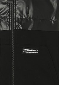 Černá zipová bunda s lesklou, vycpanou horní částí. Obsahuje přední kapsu a štítek s nápisem "KARL LAGERFELD 21 RUE ST-GUILLAUME PARIS."
