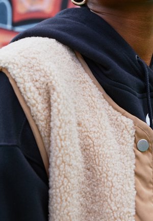 Personne portant un gilet sherpa beige par-dessus un sweat à capuche noir avec un bouton-pression argenté visible sur le gilet.