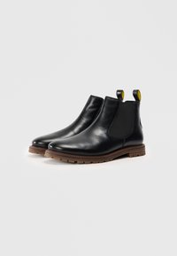 Botas de tobillo de cuero negro con paneles laterales elásticos, punta redonda y suelas de goma marrón. Presentan lengüetas amarillas para facilitar su uso.