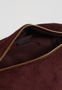 Bordowa torba z kordurowego materiału z złotym zamkiem, częściowo otwarta, ukazująca gładką podszewkę i małą czarną metkę wewnątrz.