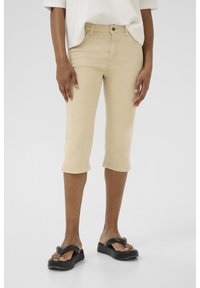 Beige cocco-housut, valmistettu denimmateriaalista, joissa on etuvetoketjukiinnitys, viisi taskua ja slim fit -muotoilu. Yhdistetty mustiin flip-flop-kenkiin.