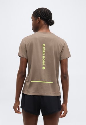 Hátraforduló nő, fonott hajjal, barna rövid ujjú ingben, amelyen sárga "BJÖRN BORG" felirat látható, és fekete rövidnadrágban.