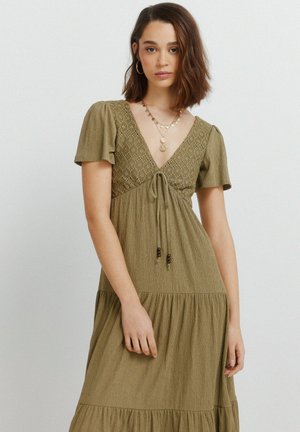 Ung kvinde med skulderlangt brunt hår iført en olivengrøn kjole med blonder og lagdelte halskæder, stående mod en helt hvid væg.