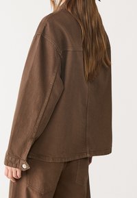 Giacca in denim marrone con vestibilità comoda, maniche lunghe e bottoni in metallo come dettagli. Presenta cuciture visibili e un tessuto strutturato.