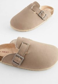 Beige läderträsko med en justerbar spänne, mjuk mockatextur och en naturligt färgad sula. Brun innerslip synlig.