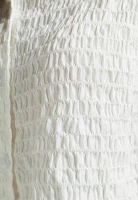 White, textured fabric with a gathered design featuring horizontal pleats. Button closure visible on the left side of the image.