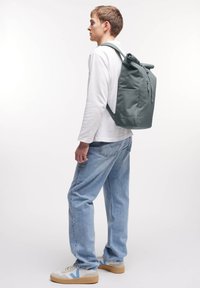 Plecak typu roll-top w stonowanym zielonym materiale, wyposażony w przednią kieszeń na zamek błyskawiczny, regulowane paski i elegancki prostokątny kształt.