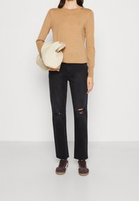Lichtbruin, aansluitend truitje, zwarte distressed jeans met knie-scheuren, en bruine high-top sneakers. Trui onder één arm gehouden.