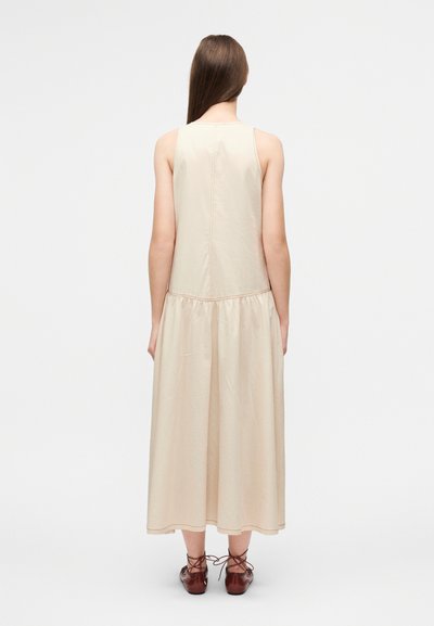 Ανοιχτό μπεζ αμάνικο φόρεμα με πιετά περιφέρεια, κατασκευασμένο από λείο ύφασμα. Διαθέτει ευθύγραμμη σιλουέτα και εμφανές κάθετο ραφές στην πλάτη.