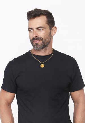 Auksinė vėrinys su apvaliu pakabuku kabantis ant plonos grandinėlės vyro, dėvinčio aptemptą juodą marškinėlį, kakle.