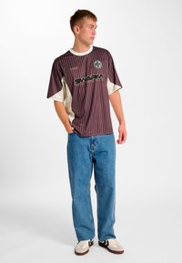 Vinrød kortærmet skjorte med striber og cremefarvet detalje samt "SWARM" logo, kombineret med lyseblå løstsiddende jeans og brune sneakers.