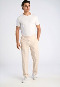 Weißes Kurzarm-T-Shirt kombiniert mit beigen Hosen mit elastischem Bund und weißen Sneakers, vor einem neutralen Hintergrund stehend.