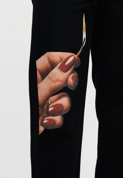 Μαύρο παντελόνι με γραφικό σχέδιο ενός χεριού που κρατά ένα σπίρτο, με μακριά, στρογγυλεμένα μανικιούρ βουργουνδίας και ρεαλιστική υφή.