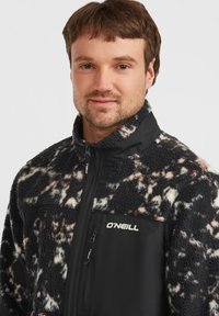 Fleece-Trainingsjacke mit einem schwarz-weißen abstrakten Muster, einem einfarbigen schwarzen Vorderteil und einer Reißverschlusstasche auf der Brust.