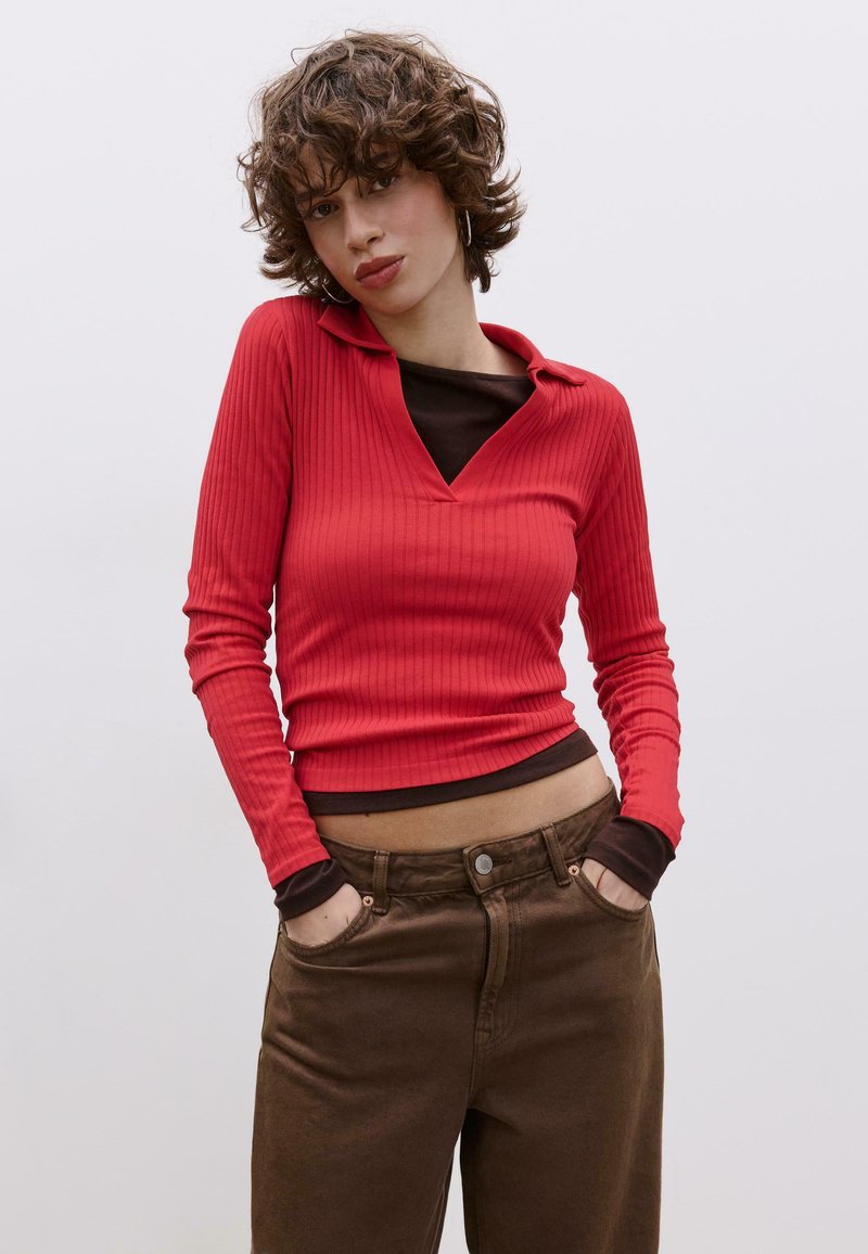 Haut rouge à manches longues côtelé avec col, superposé sur un haut marron, associé à un pantalon marron. Il présente un col en V et une coupe ajustée.