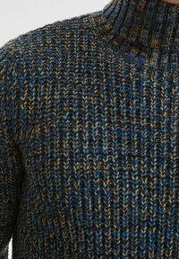 Pull en tricot avec un col haut, présentant un motif texturé aux teintes bleues, marron et dorées. Le fil épais donne une apparence chaleureuse.