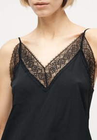 Femme portant un caraco noir orné d'une délicate bordure en dentelle florale au niveau de l'encolure et des bretelles, dévoilant le haut de la poitrine et les épaules.