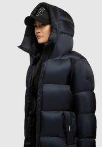 Žena nosi crnu puffer jaknu s velikom kapuljačom i crnu bejzbolsku kapu s bijelim logom, okrenuta lijevo na sivoj pozadini.