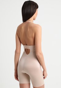 Naadloze huidskleurige catsuit met halterhalsontwerp. Heeft een open rug,  midden-dij lengte, en een discrete haaksluiting. Soepele textuur.