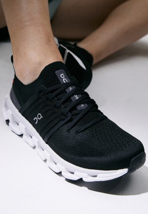 Gros plan d'une personne portant des chaussures de sport noires et blanches avec des semelles rembourrées, assise sur une surface claire.