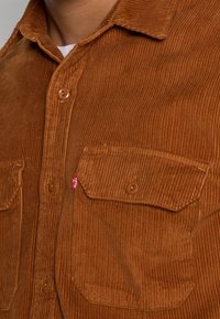 Camisa de pana en un rico marrón, con cierre de botones en la parte delantera, dos bolsillos en el pecho y una pequeña etiqueta roja. Tejido texturizado con costillas visibles.