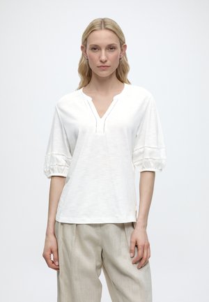 Blonde Frau steht frontal, trägt eine lockere weiße Bluse mit ellbogenlangen Puffärmeln und beige Leinenhosen.