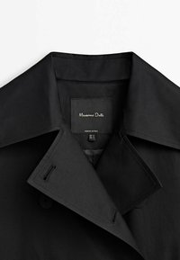 Musta trench-takki, jossa on suuri kaulus, napin yksityiskohtia ja tarra, jossa lukee "Massimo Dutti". Materiaali näyttää sileältä ja kevyeltä.