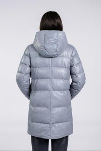 Person med mørkt hår iført en lang, lys grå, hætteklædt dunjakke, der står med ryggen til mod en ensfarvet baggrund.