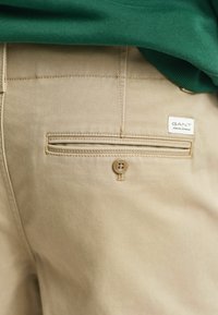 Beige cotton pants with a back pocket featuring a button closure. A white GANT label is also visible near the pocket.
