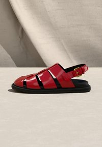 Κόκκινο γυαλιστερό δερμάτινο σανδάλι ψαρά με μαύρη σόλα και χρυσή αγκράφα, τοποθετημένο σε ουδέτερη υφή επιφάνεια με απαλή σκιά.