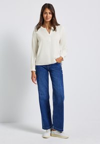 Femme se tenant avec une main dans la poche, portant un blouse longue à manches blanches, un jean large bleu et des baskets blanches, sur fond uni.